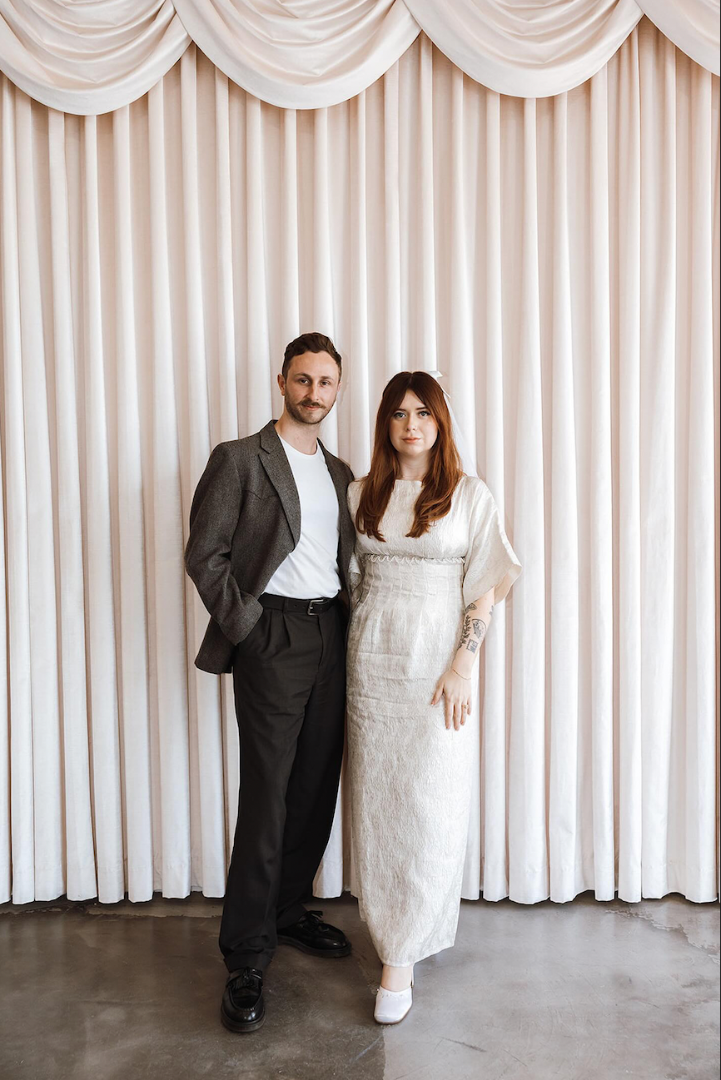 A man in a vintage suit and a woman in a white, vintage bridal dress standing next to each other with a white curtain as the backdrop.