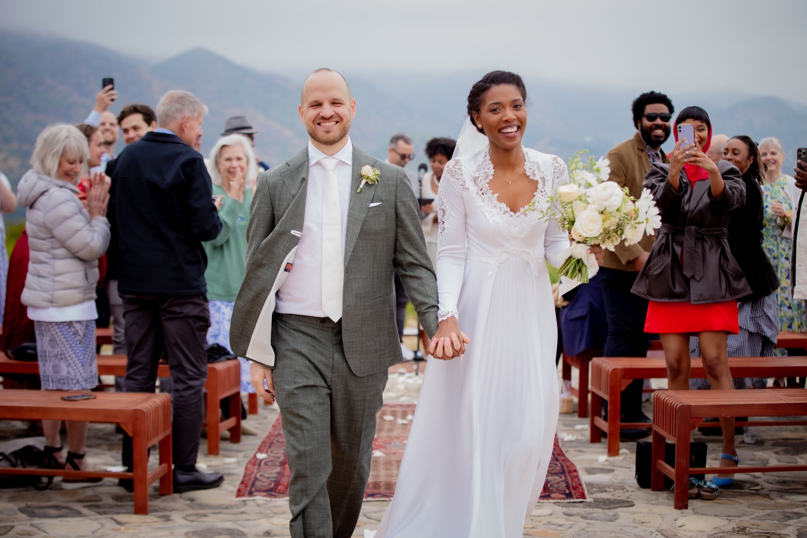 A white groom and African-American bride walking back down the aisle with friends and family cheering and taking pictures. 