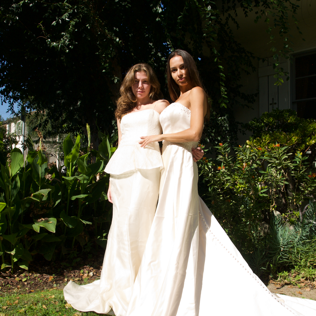 Two girls in white wedding dresses posing in front of green bushes and trees.