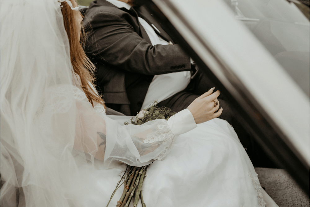 A close up shot of the bride and groom sitting in a car. The groom is driving, and the bride is holding a bouquet of flowers with her wedding ring displayed on her finger.
